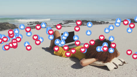 Animation-of-red-heart-love-and-like-digital-icons-over-woman-using-laptop-on-beach