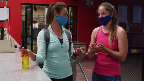 Two-caucasian-women-wearing-face-masks-talking-in-foyer-of-gym-and-disinfecting-hands