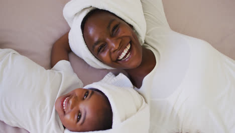 Portrait-of-african-american-mother-and-daughter-wearing-towels-lying-on-bed-and-smiling