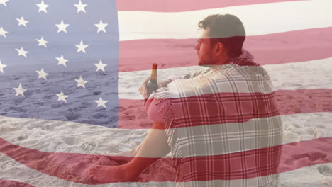 Animation-of-american-flag-waving-over-man-wrapped-in-blanket-drinking-beer-on-beach