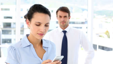 Geschäftsfrau,-Die-Mit-Einem-Kollegen-Hinter-Ihr-Eine-SMS-Auf-Ihrem-Smartphone-Schreibt
