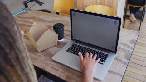 Mixed-race-man-with-dreadlocks-sitting-in-cafe-with-sandwich-and-laptop-with-copy-space-on-screen