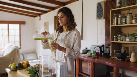 Happy-mixed-race-woman-pouring-health-drink-standing-in-cottage-kitchen-smiling