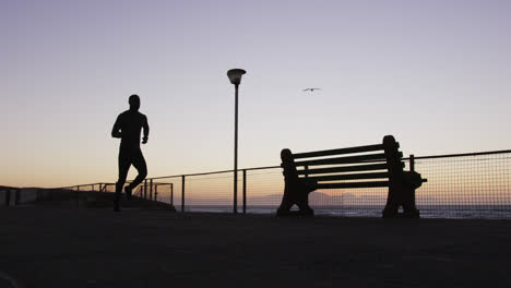 African-american-man-exercising-outdoors,-running-by-seaside-in-the-evening