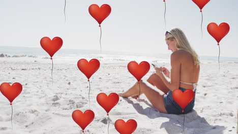 Animation-of-red-heart-love-balloons-digital-icons-over-woman-using-smartphone-on-beach