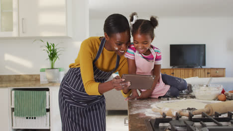 Happy-african-american-mother-and-daughter-cooking-and-looking-at-digital-tablet-in-kitchen