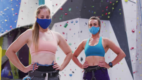 Portrait-of-two-caucasian-women-wearing-face-masks-standing-at-indoor-climbing-wall