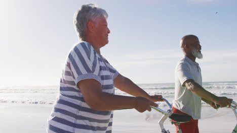 Smiling-senior-african-american-couple-walking-with-bicycles-at-the-beach