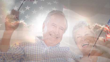Animation-of-glowing-light-over-portrait-of-happy-senior-couple-waving-american-flags