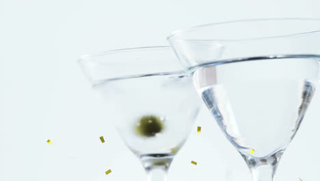 Golden-confetti-falling-over-olives-falling-into-cocktail-glasses-against-grey-background