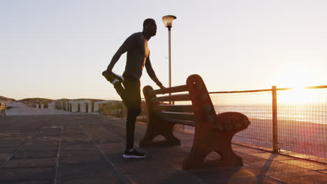 Focused-african-american-man-stretching-by-bench,-exercising-outdoors-by-seaside-at-sunset