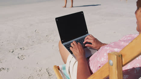 Mixed-race-senior-woman-sitting-on-sunbed-and-using-a-laptop-at-the-beach