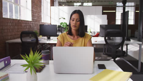 Mixed-race-businesswoman-sitting-at-desk-using-laptop-looking-ahead