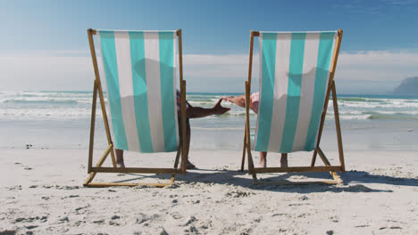 Älteres-Afroamerikanisches-Paar-Sitzt-Auf-Sonnenliegen-Und-Hält-Händchen-Am-Strand