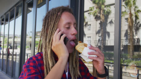 Mixed-race-man-with-dreadlocks-walking-on-the-street-eating-sandwich-and-talking-on-smartphone