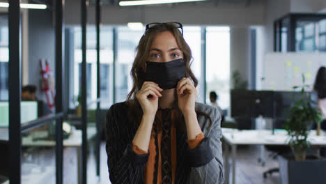 Portrait-of-caucasian-businesswoman-adjusting-face-mask-standing-in-office-and-looking-to-camera