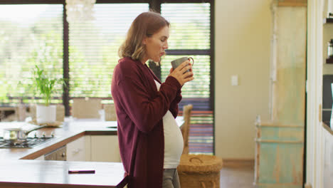 Caucasian-pregnant-woman-standing-in-kitchen,-touching-belly-and-drinking-tea