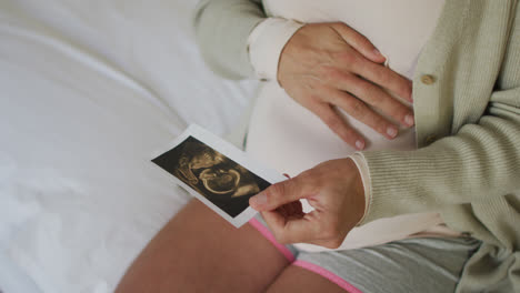 Manos-De-Una-Mujer-Embarazada-Caucásica-Sentada-En-La-Cama,-Tocando-El-Vientre-Y-Mirando-La-Foto-De-Ustrasound