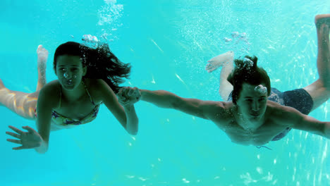 Happy-couple-holding-hands-underwater-in-swimming-pool-and-waving