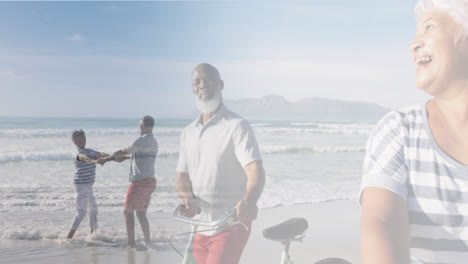 Composite-of-happy-diverse-senior-couple-walking-with-bikes,and-dancing-on-beach