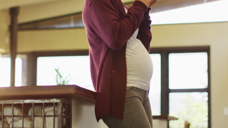 Midsection-of-caucasian-pregnant-woman-standing-in-kitchen,-touching-belly-and-drinking-tea