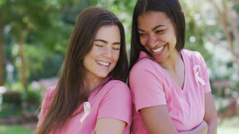 Two-happy-diverse-women-in-pink-t-shirts-and-cancer-ribbons,-talking-and-laughing-in-park