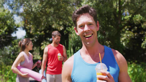 Un-Hombre-Caucásico-Sonriente-Bebiendo-Bebidas-Saludables,-Con-Un-Grupo-Diverso-Hablando-Después-De-Yoga-En-Un-Parque-Soleado