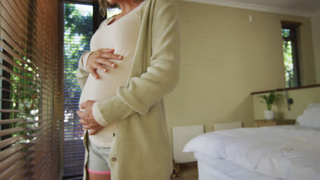 Midsection-of-caucasian-pregnant-woman-standing-at-window-and-touching-belly