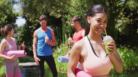 Smiling-biracial-woman-drinking-health-drink,-with-diverse-group-talking-after-yoga-in-sunny-park