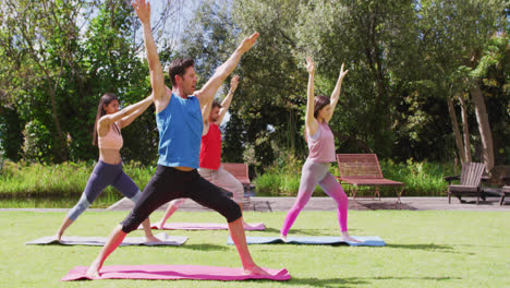 Grupo-Diverso-Practicando-Yoga-Con-Un-Instructor-Masculino,-Parado-Sobre-Colchonetas-En-Un-Parque-Soleado