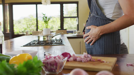 Midsection-of-caucasian-pregnant-woman-wearing-apron,-touching-belly-in-kitchen