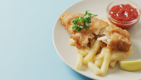 Video-close-up-of-fish-and-chips,-parsley-and-bowl-of-ketchup-on-plate,-with-copy-space-on-blue