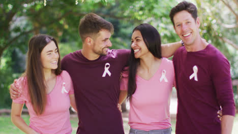 Portrait-of-happy,-diverse-group-of-men-and-women-wearing-cancer-awareness-ribbons-embracing-in-park