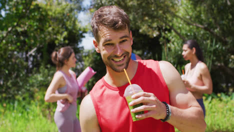 Smiling-caucasian-man-with-health-drink,-and-diverse-group-talking-after-yoga-in-sunny-park