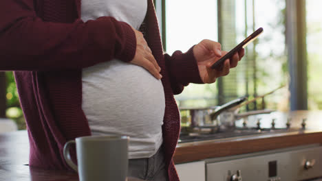 Sección-Media-De-Una-Mujer-Embarazada-Caucásica-Tocando-El-Vientre-Y-Usando-Un-Teléfono-Inteligente