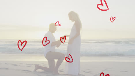 Animation-of-red-hearts-over-caucasian-man-proposing-on-beach