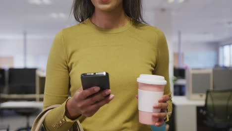 Biracial-businesswoman-smiling-and-walking-with-smartphone-in-office