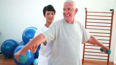 Feliz-Fisioterapeuta-Ayudando-A-Un-Paciente-Anciano-A-Levantar-Pesas-De-Mano
