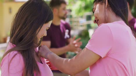 Two-happy-diverse-women-one-pinning-breast-cancer-ribbon-to-t-shirt,-talking-and-laughing-in-park