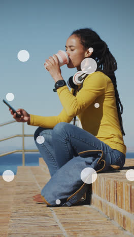 Weiße-Flecken-Auf-Einer-Afroamerikanischen-Frau,-Die-Auf-Der-Promenade-Kaffee-Trinkt-Und-Ihr-Smartphone-Benutzt