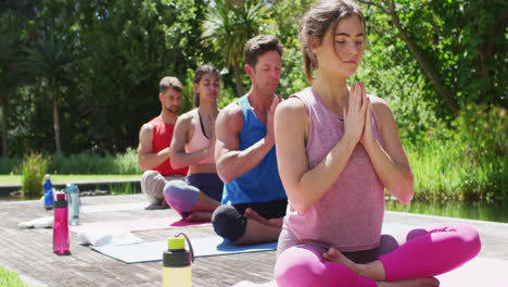 Diverse-male-and-female-group-practicing-yoga-sitting-in-a-row-with-eyes-closed-in-sunny-park