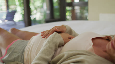 Midsection-of-caucasian-pregnant-woman-touching-belly-and-resting-on-bed