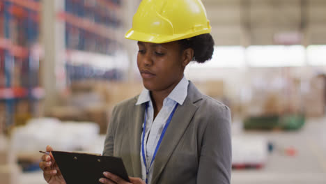 Portrait-of-african-american-female-worker-wearing-helmet-and-smiling-in-warehouse