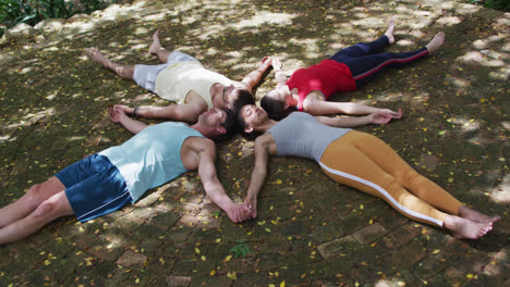 Diverse-group-of-men-and-women-lying-on-backs-touching-heads,-holding-hands-with-eyes-closed-in-park