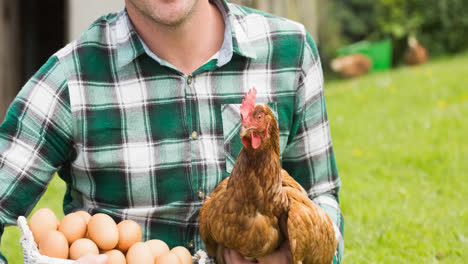 Smiling-caucasian-male-farmer-holding-free-range-hen-and-eggs-on-farmyard