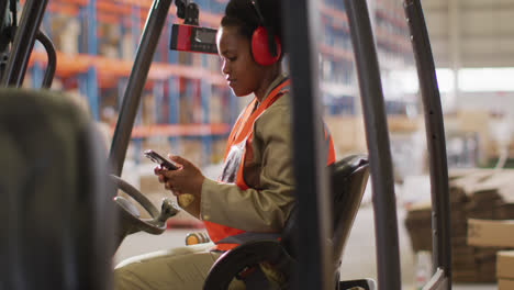 African-american-female-worker-wearing-safety-suit-and-using-smartphone-in-warehouse