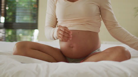 Midsection-of-caucasian-pregnant-woman-sitting-on-bed-and-putting-headphones-on-belly