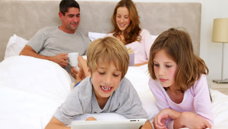 Kinder,-Die-Im-Bett-Ein-Tablet-Benutzen,-Während-Ihre-Eltern-Hinter-Ihnen-Plaudern