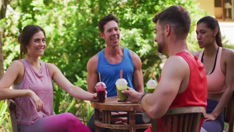 Smiling-diverse-group-sitting-at-table-with-health-drinks,-talking-after-yoga-in-sunny-park