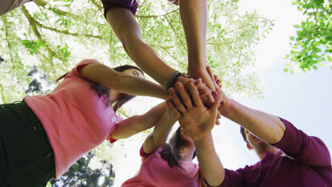 Diverse-group-of-men-and-women-wearing-breast-cancer-ribbons,-stacking-hands-and-laughing-in-park